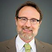 A headshot of a person with glasses, a beard, and mustache, wearing a brown suit and a green tie, smiling against a gray background.