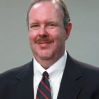 Headshot of a person with short hair and a mustache, wearing a suit, white shirt, and striped tie.