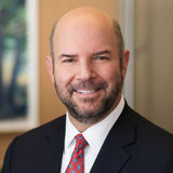 A professional headshot of a person with a beard, wearing a suit and red tie, smiling, with a blurred background.