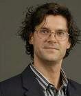 Headshot of a person with glasses, wavy hair, wearing a striped shirt and dark jacket, against a neutral background.