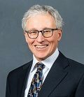 Headshot of a person wearing glasses, a dark suit, a white shirt, and a patterned tie, smiling against a gray background.