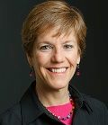 Headshot of a smiling person with short, light brown hair, wearing a black jacket and pink top, on a dark background.