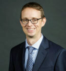 A headshot of a person wearing glasses, a suit, and a tie, with a neutral background.