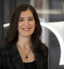 Headshot of a smiling person with long dark hair, wearing a necklace and a dark blazer, standing against a blurred background.