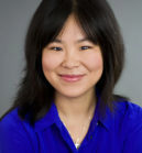 Headshot of a person with long dark hair, wearing a blue shirt, smiling slightly, against a gray background.